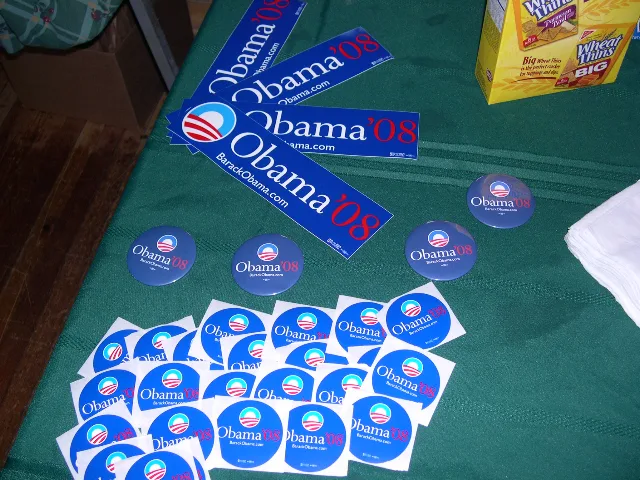 On a table, different shapes of blue Obama ‘08 stickers and buttons are displayed. In the background is a Wheat Thins box. 