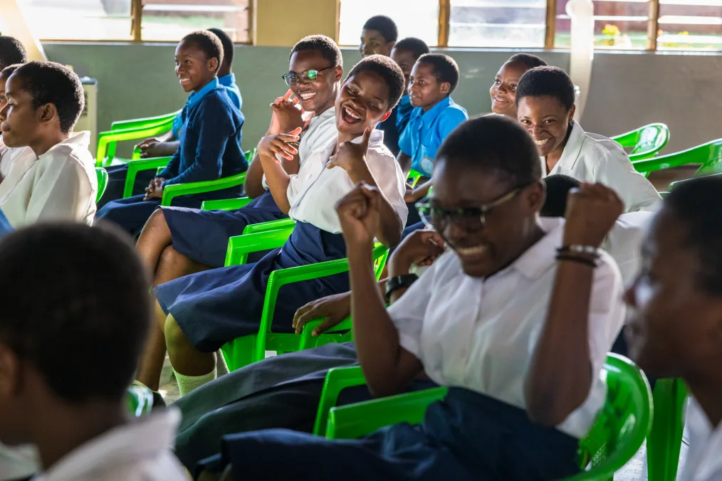 A group of young girls sit on green chairs in a classroom. They are a range of medium and deep skin tones and they are wearing white and blue uniforms. Many girls wear glasses and have low haircuts. 