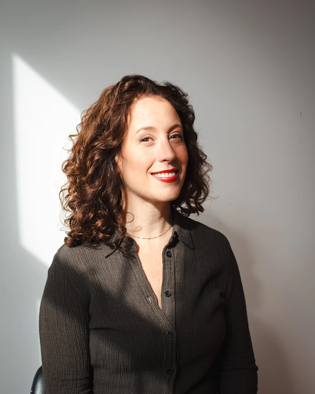 The image is a photographed portrait of Aliza Nisenbaum. Aliza is sitting in a black chair that is partially obscured in a room with a white wall. Alize has an olive complexion and long dark curly hair. She is smiling at the camera. She is wearing a small dark necklace and a dark colored button-down shirt.