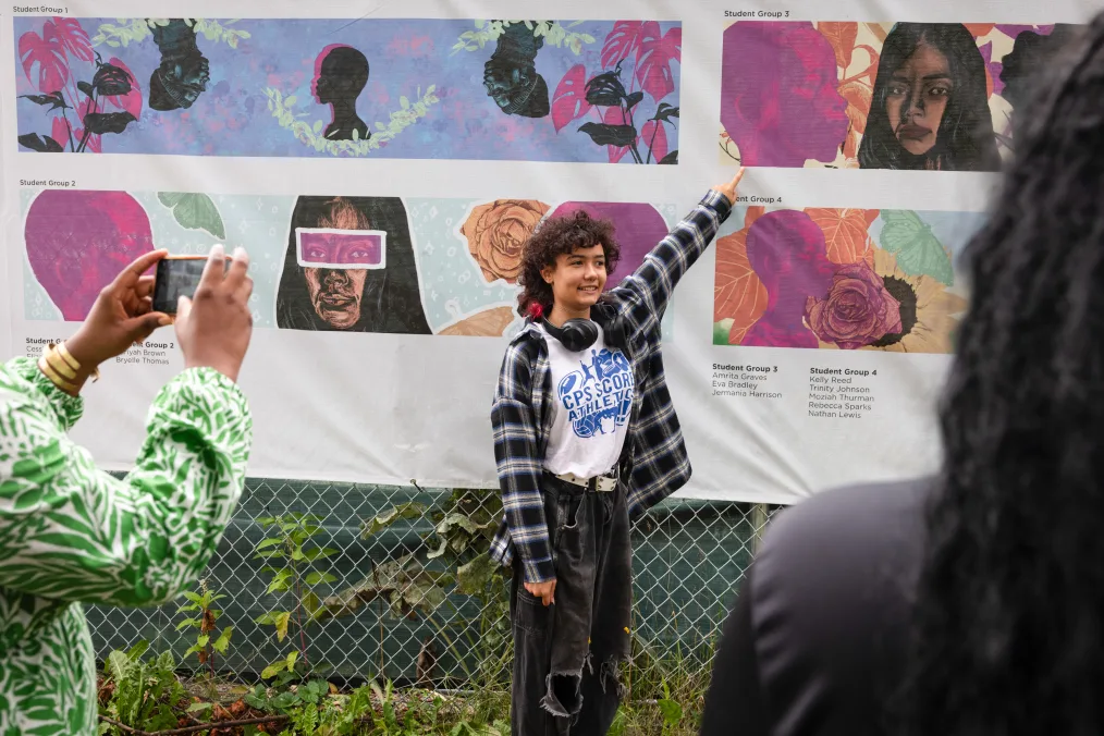 Eva Bradley, a young woman with a light medium skin tone, stands and points at a mural at the site of the Obama Presidential Center. Eva is wearing a flannel t-shirt and has curly brown hair with pink tips. The mural in the background features illustrations of young people.