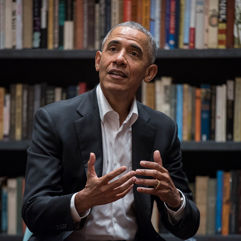 A close shot of President Obama speaking at a Chicago Public Library branch. Books on shelves are in the background.  President Obama has a medium deep skin complexion and gray and black closely cut hair. He is wearing a dark colored suit. 