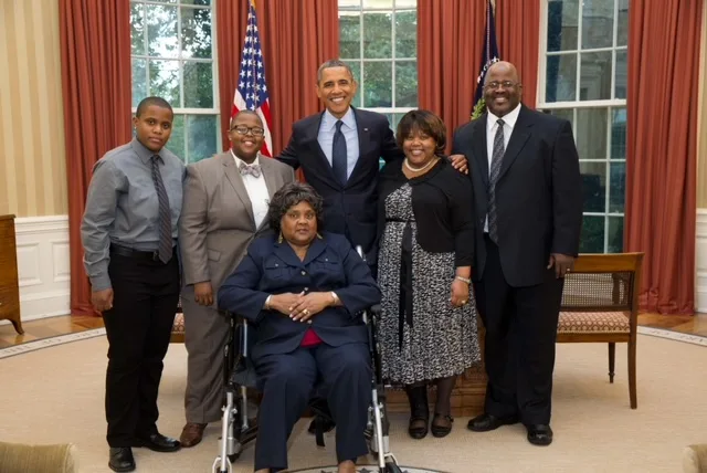 An image on Ashlee Davis with President Obama posing with her family in the Oval Office. Ashlee has a medium deep complexion and shortly cropped black hair. She is wearing a taupe suit with a taupe bow tie.