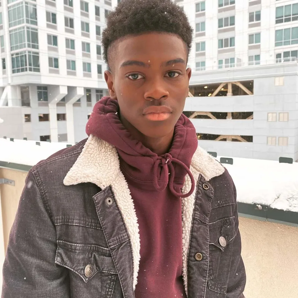 A  boy with a medium-deep skin tone takes a picture with snow and buildings in the background