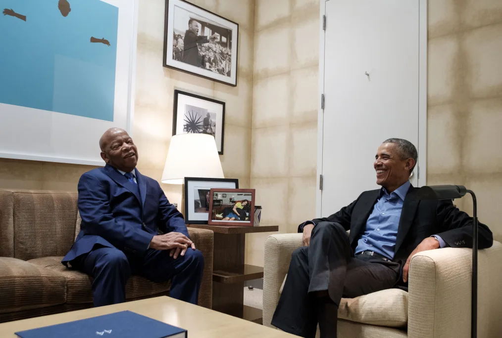 Representative John Lewis and President Barack Obama are sitting in a room together laughing in the middle of a conversation. Representative Lewis is sitting on a couch on the left-hand side of the image with his hands placed on his left knee. He is an older, brown-skinned man with a bald head. He is wearing a blue suit, with a white button-down shirt, and a blue tie. The couch Rep. Lewis is sitting on, is the color brown. President Obama is sitting on the right side of the image in a beige chair. He is wearing a black suit with a blue button-down shirt. His arms are placed on the side of the chair. In the room of the image is a beige coffee table with a large blue book on top of it. On the front of the book is the number, “44.” The room has beige walls with pictures on the left side. On the wall are three pictures: the largest picture features a blue background and the figure of a man’s arms and head; the second largest picture is an image of Dr. Martin Luther King Jr. speaking during the March on Washington; the smallest picture is indistinguishable. In the room there is a small brown side table that sits between Rep. John Lewis and President Barack Obama. On the side table is a table lamp with a white lamp shade and two framed pictures.