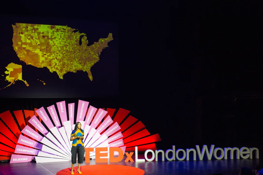 Vidhya Ramalingam stands on a stage and speaks at a TedxLondon Event. She has a medium skin tone and is wearing a blue and yellow patterned top. On the stage behind her is a gold map of the United States, a graphic that reads, “TEDxLondon”
