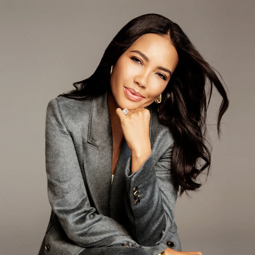 A photographic portrait of woman with medium skin tone and long black hair posing for the camera with her chin resting on her left hand and her head tilted to the side. She wears a gray suit jacket, earrings and a large ring. 