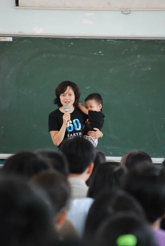 A woman with a light skin tone and short black hair stands in front of a chalkboard while holding a microphone. In her other hand, she holds a baby with a light skin tone. They stand together in front of a large crowd of various people 