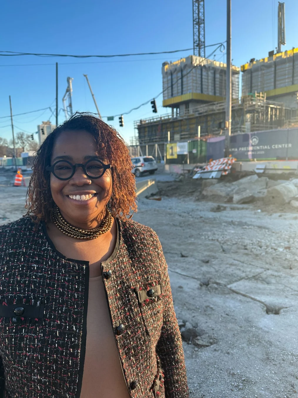 Crystal Moten, PhD, a Black woman with a brown locs and a medium deep skin tone, smiles at the camera. In the background is construction at the site of the Obama Presidential Center. She is wearing black circular glasses, a brown beaded necklace, and a black tweed jacket.