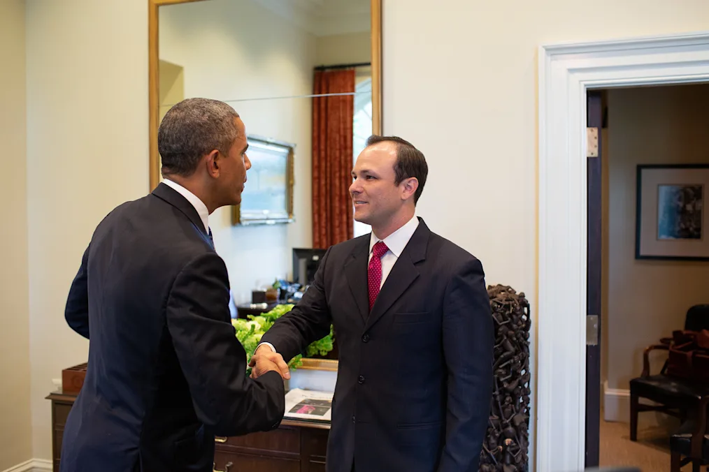 An image of Charlie Galbraith shaking hands with President Barack Obama. President Obama is facing away from the camera. In the background of the picture is a large mirror with a gold trim. Underneath the mirror is a dark brown desk with gold pulls. Next to the desk is an abstract statue composed of small figures of humans. There is an open door to the right of the picture. Through the doorway is a small chair and a picture hanging on a white wall. Charlie has an olive complexion and short dark hair. He is wearing a dark colored suit and a pink tie with a white shirt underneath.