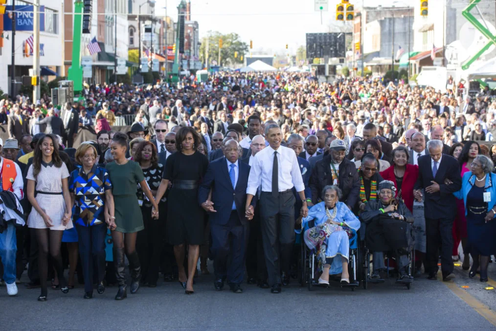 President Obama, his family, and thousands of people, and John Lewis cross the Edmund Pettus Bridge hand in hand.