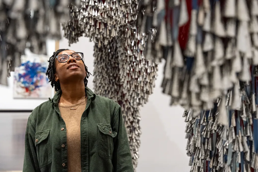 An image of a person looking at Marie Watt's installation Sky Dances Light. The person has a deep skin tone and short locs. They are wearing glasses, a green army jacket, and a beige shirt. They are looking up at the installation. The installation is made up of tin jingles hanging from the ceiling. The jingles are silver cones. There are thousands of them. In between the jingles are straps of pink and blue fabric.