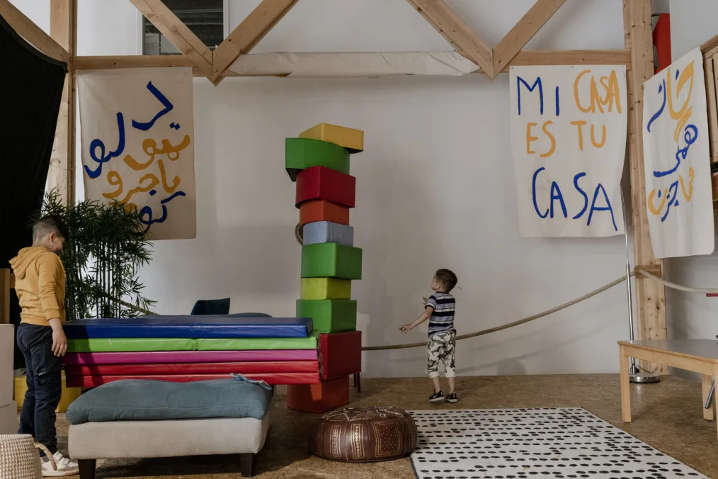 There are two young boys with light skin tones standing in a room with white walls. There is a wooden structure built slightly in front of the wall with three white posters with yellow and blue writing. There are rainbow-colored blocks stacked up and mats stacked up on the left-hand side of the photo where there is also a plant. There is a small beige stool with a blue cushion next to a small brown patterned floor seat. 
