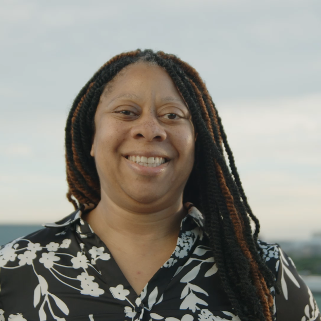 The image is a gif of Melanie Moore smiling. Melanie has a light medium deep complexion, dark brown eyes, and long braided twists in her hair that are the colors black and brown. She is wearing a black collared blouse with white flowers on it. She is standing outside in the daytime with the Chicago skyline in the background.