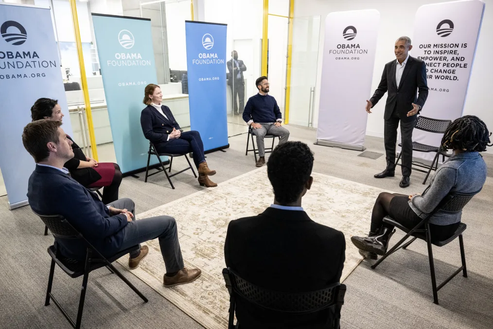 A photo of President Obama and a circle group of middle aged men and women. They are in a small office space, that has see in glass on the left. On the floor is a yellow/beige carpet with brown designs. Each person sits in a light black chair except President Obama who is standing and saying something. On their right are a set of spread out Obama Foundation Posters. Two men and women sit with light skin are sitting in the left of the photo in formal attire. On the right is one man and woman with medium to deep skin tones in formal attire. There faces are not visible to the camera.