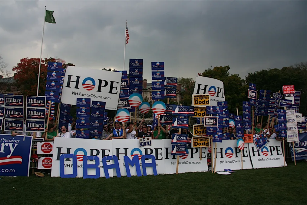 A group of people hold up dozens of large political signs, including ones that read "HOPE," "Obama," "Hillary, "I believe," "Firefighters for Dodd." Large blue cut-out letters spell "Obama" in the front. Behind them are trees and two flags flying on flagpoles.   