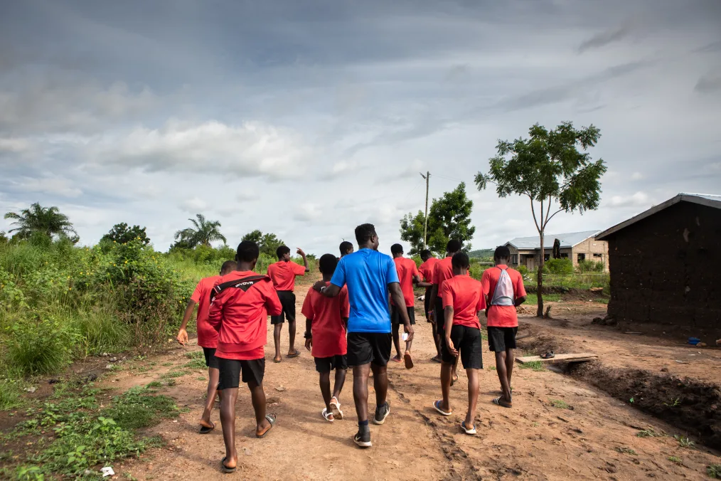 A group of young men wearing red shirts walking with a man wearing a blue shirt in, all medium brown skin 