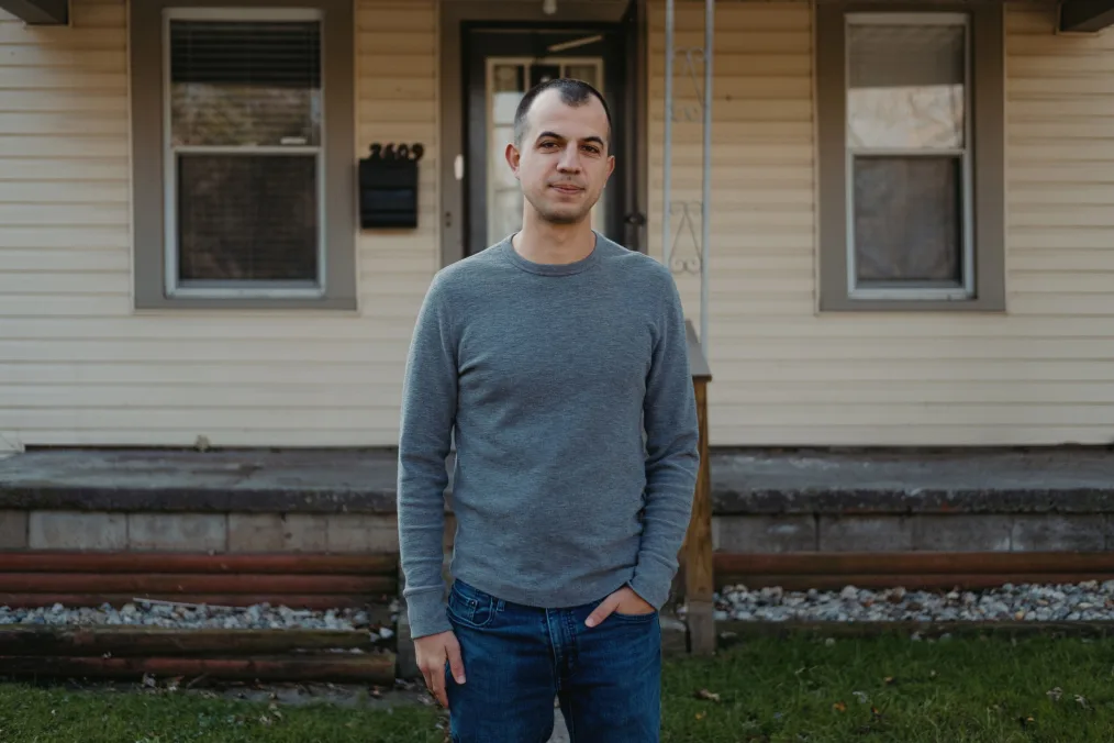 A man in a gray long sleeve shirt and jeans stands in front of a home in Toledo, Ohio. He has one hand in his pocket and is smirking directly at us.