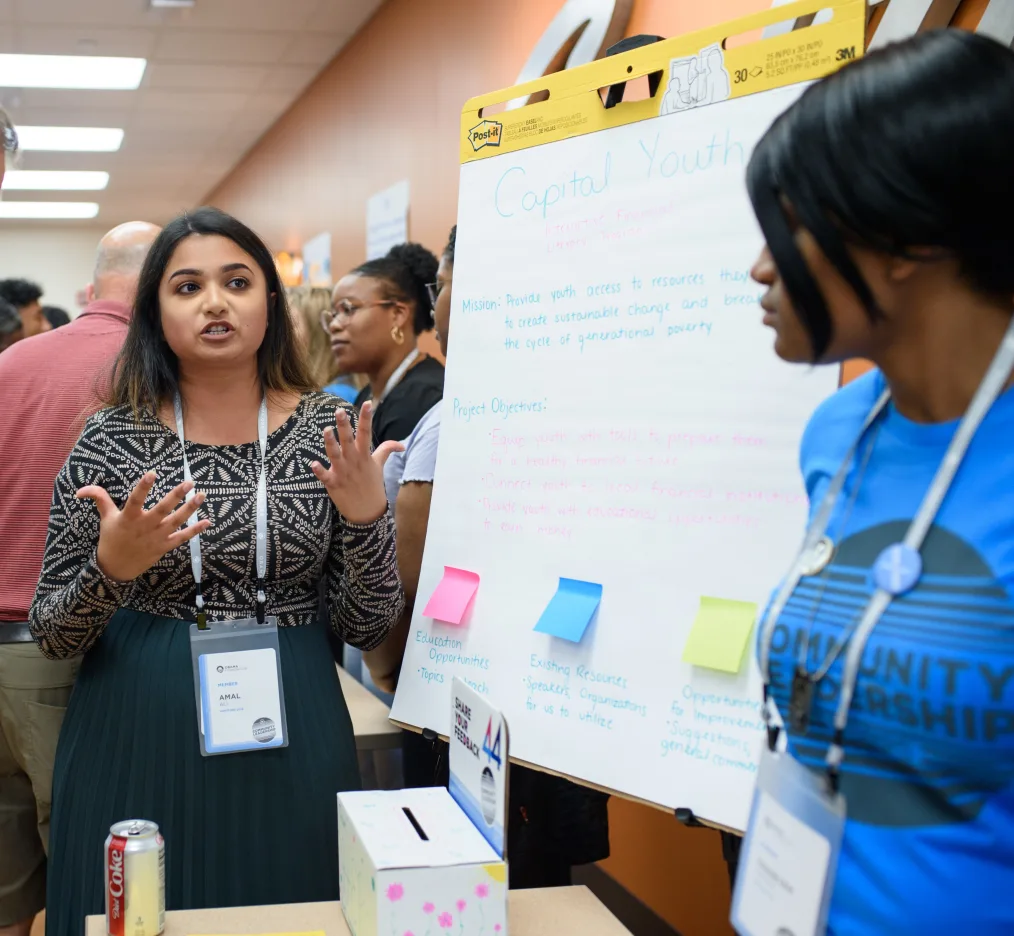 A woman with a medium skin tone with a teal skirt, black and tan patterned shirt, and an identification tag on a lanyard around her neck, speaks using hand gestures to another woman of a medium-deep skin tone wearing and blue shirt with black letters as they stand on opposite sides of a large notepad, leaning up against a canvas, with blue and pink writing and a blue, pink, and yellow sticky note. There are other people moving around the room in the background. 