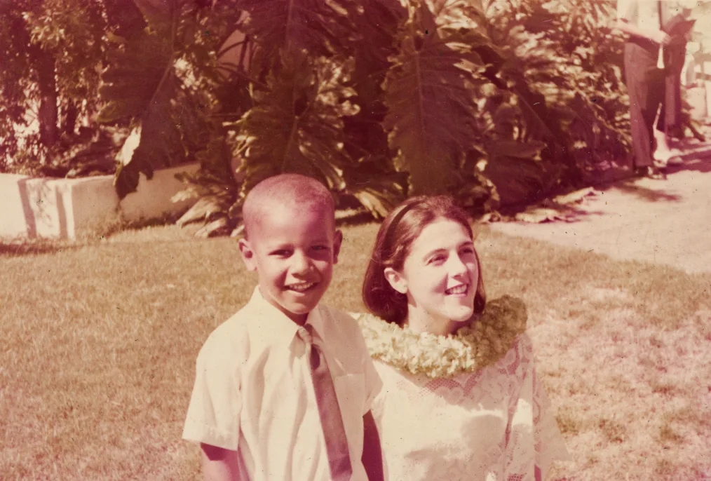A young Barack Obama wearing a short sleeve shirt and tie poses for the camera next to his mother, Ann Dunham, who wears a white lace top and a necklace of flowers. They stand in a grassy space with foliage behind them.