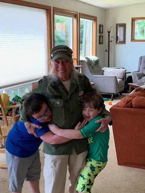 An older woman wearing a hat hugs two little kids, smiles to camera.