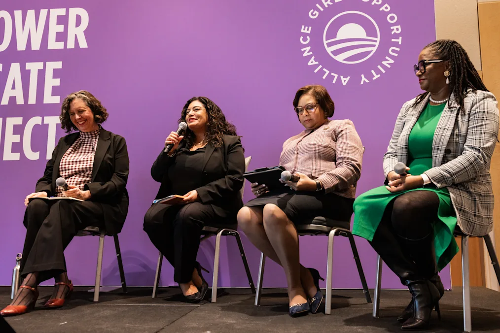 A group of women with a range of light to dark skin tones sit on a panel. In the background, a purple banner reads, “Empower, Educate, Connect” and “Girls Opportunity Alliance.”