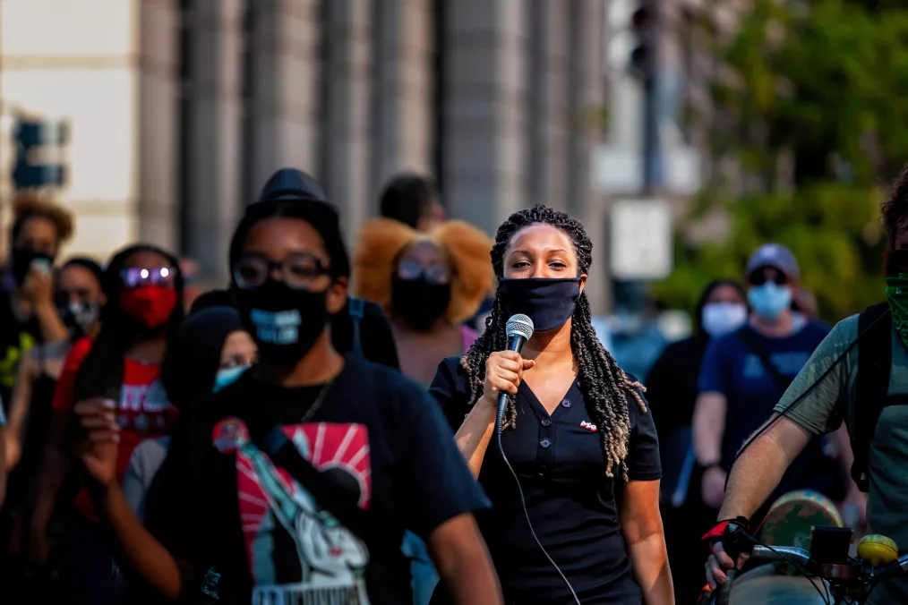 A woman with medium-dark skin tone, long hair in tightly coiled braids, and a black facemask holds a microphone. Around her people appear to walk down the same direction. 