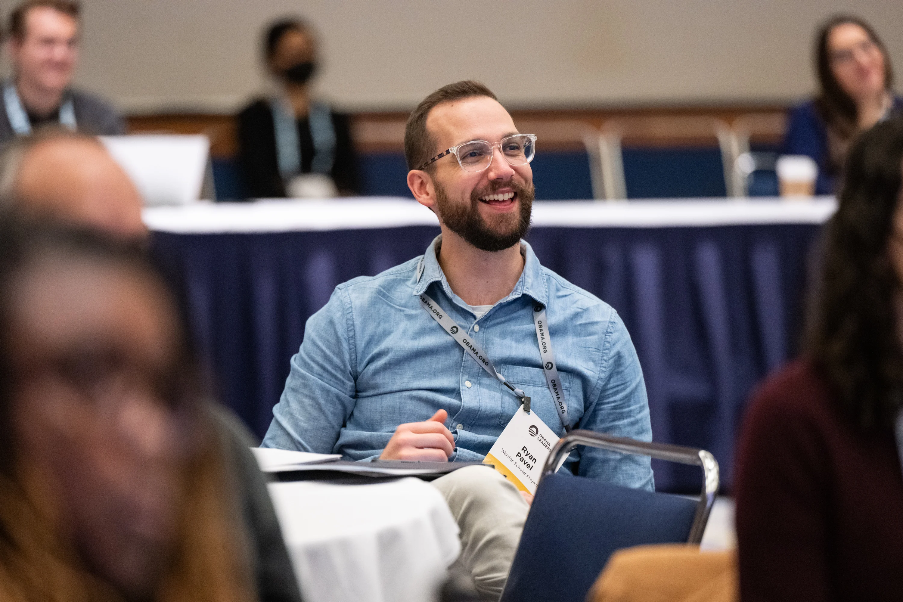 Ryan Pavel, a man with a light skin tone, wears a lanyard with his name as he sits in a conference room behind a table. He is wearing glasses and a denim shirt.