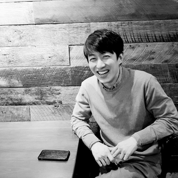 In a black and white photo, Joseph Lim, an Asian man, smiles as he sits at a table.
