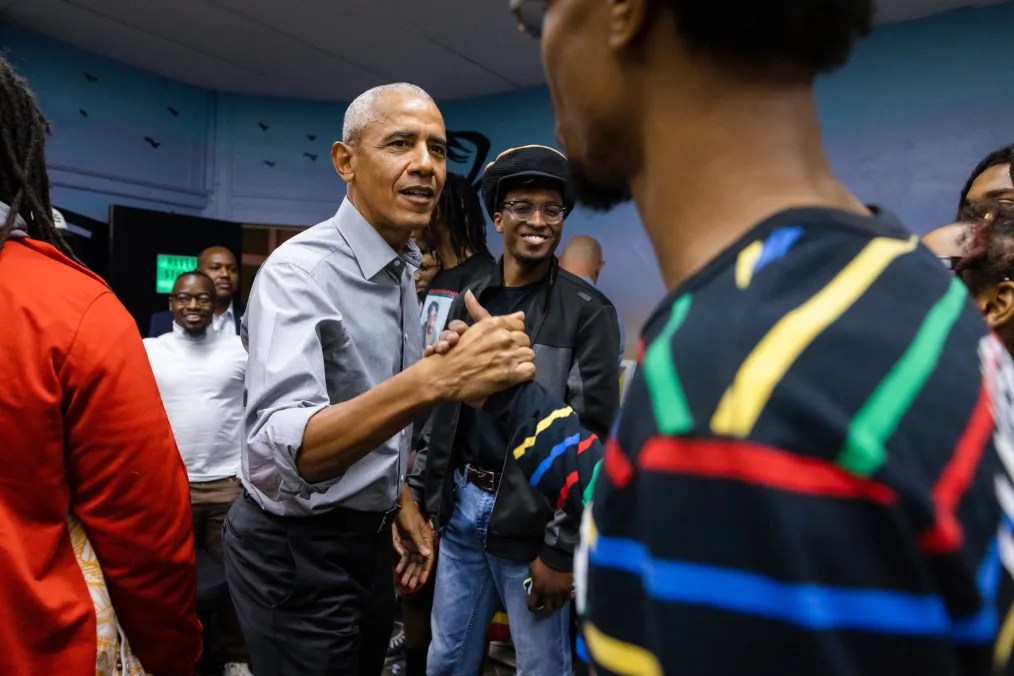 President Obama shakes the hand of a Black man with a deep skin tone and striped black shirt. Several others stand in the background.