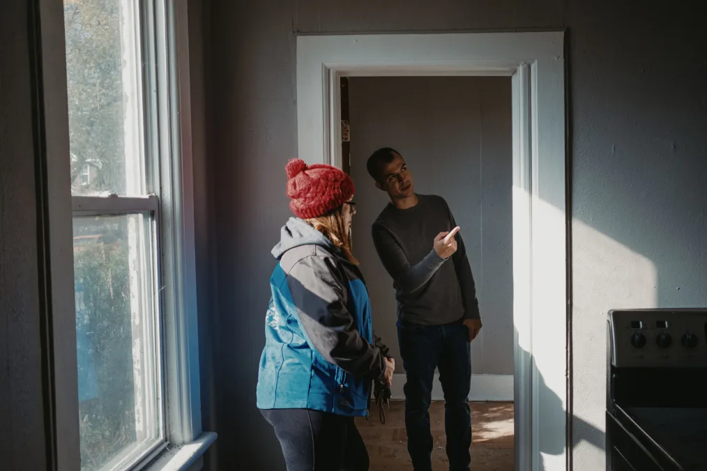 A man and woman inside of a house. The man stands under a white door frame leading to the room the woman is in. Both rooms have grey paint. The womans room on the left has a window and on the right close to the door the man is standing under is a white stove. The woman is wearing layers of clothing. A red ski cap, grey and blue jacket and jeans. She has blonde hair, light skin, and black glasses. The man in the door has short hair, a windows peak hairline, medium-light skin with warm undertones, a thin sweater, and jeans on. He is explaining something to the woman. 