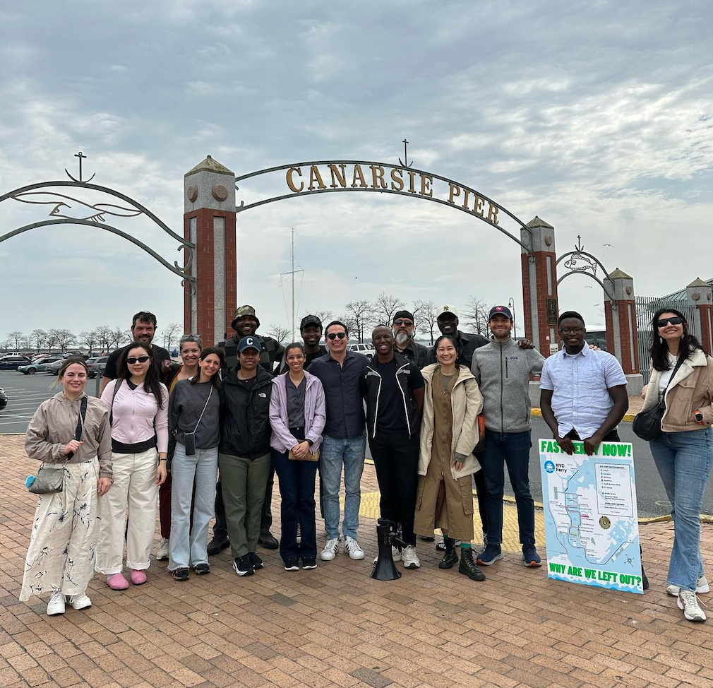 Jibreel Jalloh, a male with a dark skin tone, stands in the center of the picture wearing black pants and a black sweater, standing alongside other individuals from The Obama Scholars Program during a site visit to Canarsie Pier. All are a range of light to dark skin tones. 