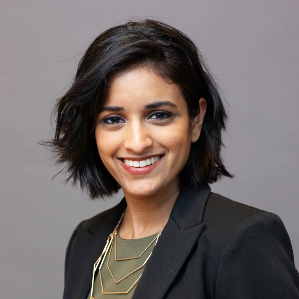 A headshot of woman with medium skin tone and wavy, black, chin-length hair smiling for the camera. She wears several geometric necklaces, and black suit jacket over a forest-green top.