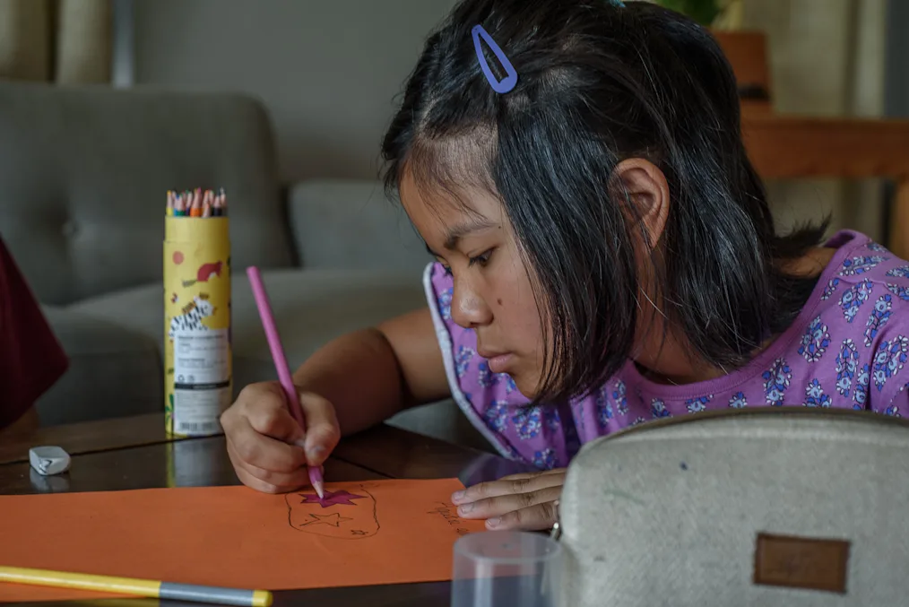 Dechen, a young Nepali girl with a medium skin tone, draws stars on paper with a yellow case of colored pencils. She is wearing a purple dress, and her chin-length black hair is held with a purple clip.  
