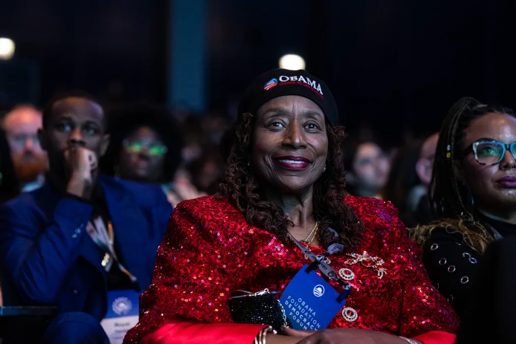 A Black woman with a deep skin tone smiles at the camera. She is wearing a red sequin top and a hat that reads, “Obama, change we can believe in.” A blue lanyard on her neck reads, “Obama Foundation Democracy Forum.” 