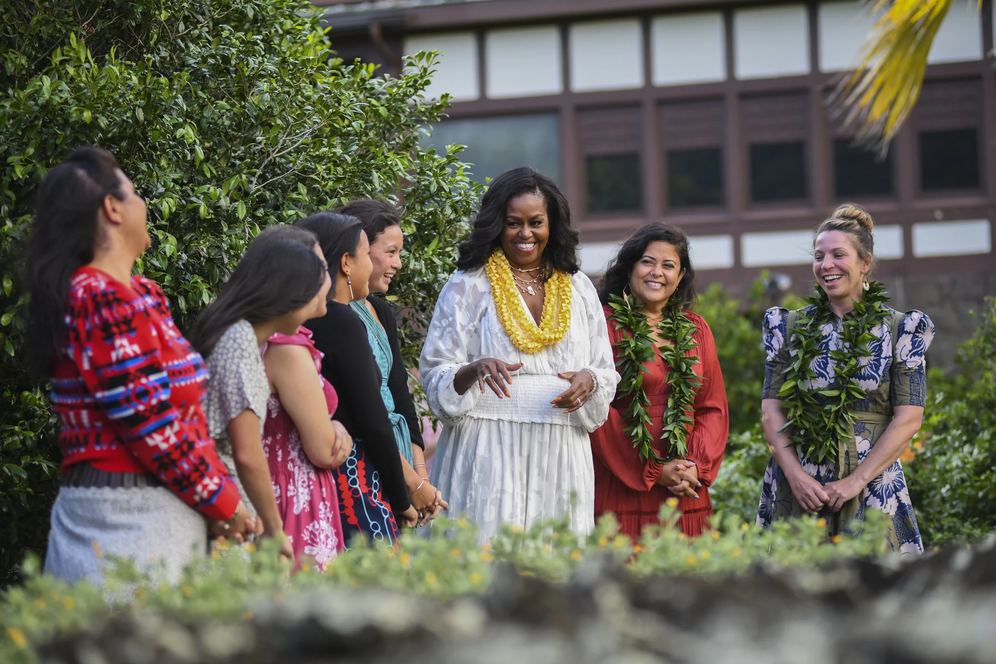 Michelle Obama enjoys a conversation with a group of women. Most of the woman have island or latin facial features. One of the woman on the right has light skin tones and blonde and brown hair tied in a bun. Most if not all are wearing a traditional dress, possibly Hawaiian. Michelle Obama is wearing identical yellow hawaiian neclaces and a white traditional dress. The other womans dresses are a variance colors. The two women on the right have neclaces of a green flat leaf plant. They are standing in a curved line outside infront of well trimmed tall bushes. In the background is a huge building with most squre based arcitechture. It is burnt dark red and white.  