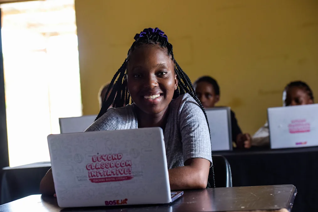 A young girl, Mosa, sits at a classroom desk with a laptop branded with “Beyond the Classroom Initiative” and two girls sitting behind her. She wears a gray blouse and has her braided hair tied up with a purple hair-tie