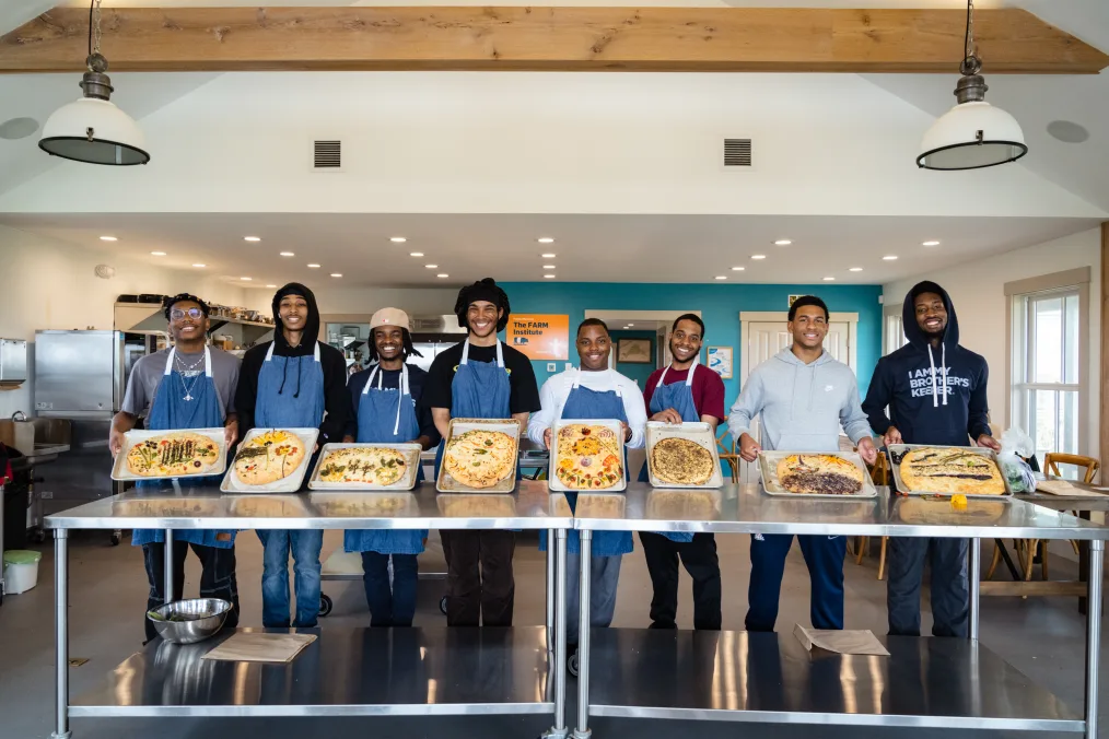 Eight Black young men with a range of medium and deep skin tones stand behind a table and hold up the focaccia bread they baked. Six of the eight are wearing blue aprons. All are standing in a kitchen.