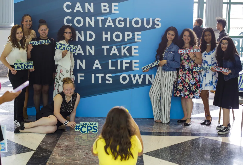 Young girls and women ranging to light to deep medium skin tone, stands infront of a large blue sign with text while holding smaller signs that reads "Chicago" , "Hopeful", and "Community Leader" as someone take their picture 