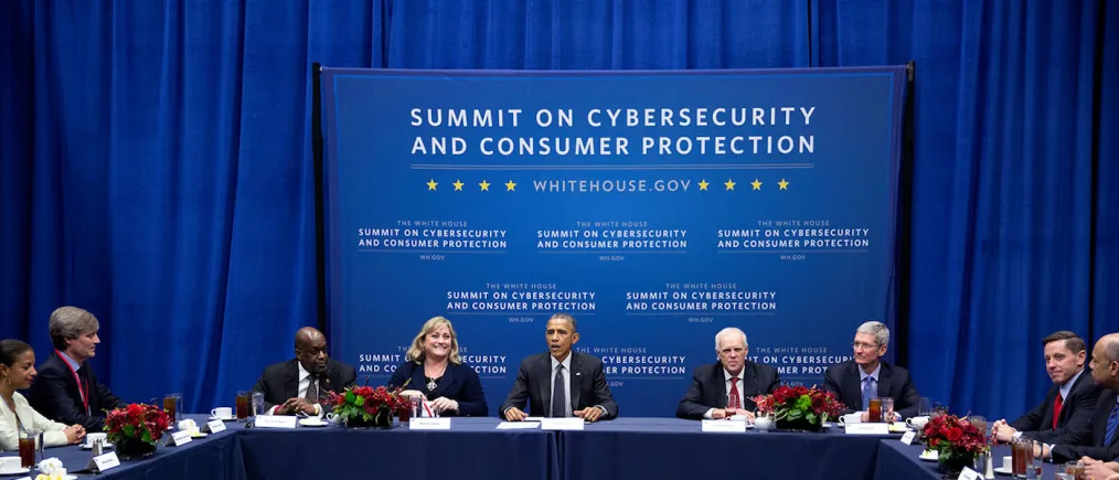 President Obama sits at a table with people of various skin tones. The background features a backdrop that reads ¨summit on cybersecurity and consumer protection¨