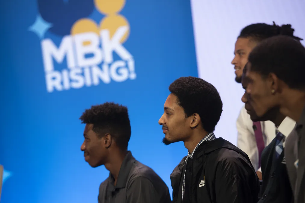 A group of young black men of various skin tones look toward the left out of frame. There is a blue graphic with ¨MBK Rising!¨ in the background.