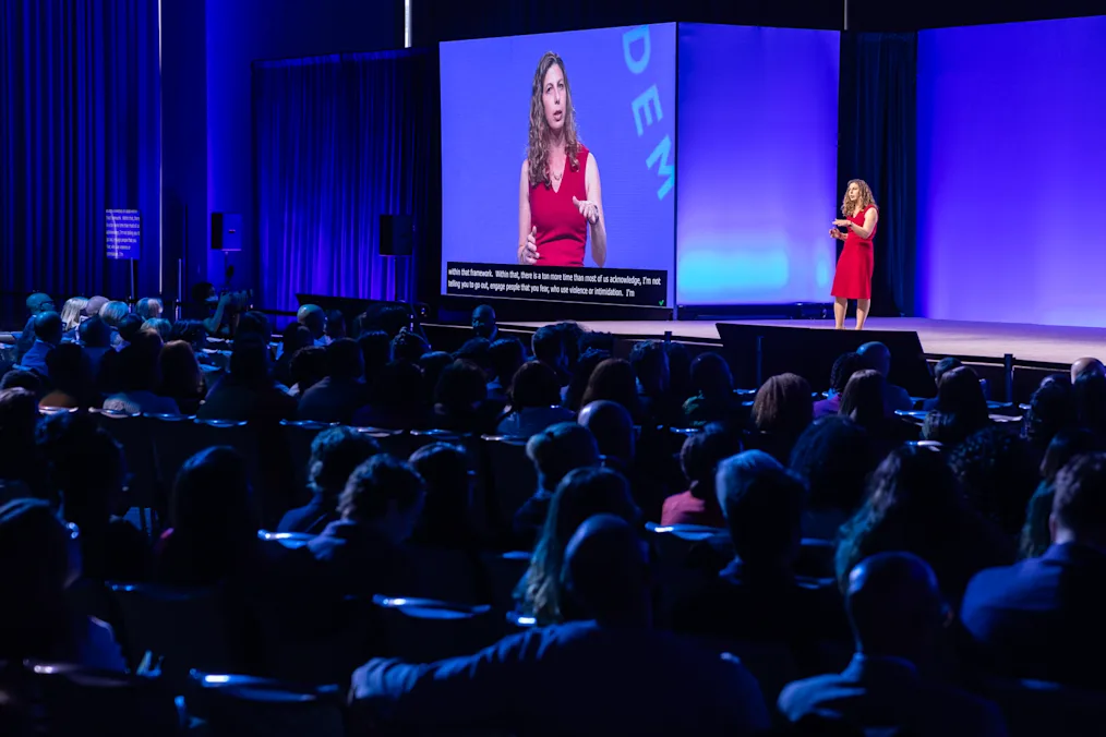 Dr. Rachel Kleinfeld, a woman with a light skin tone and curly black hair, speaks to an audience at the 2024 Democracy Forum. She is wearing a red dress. 