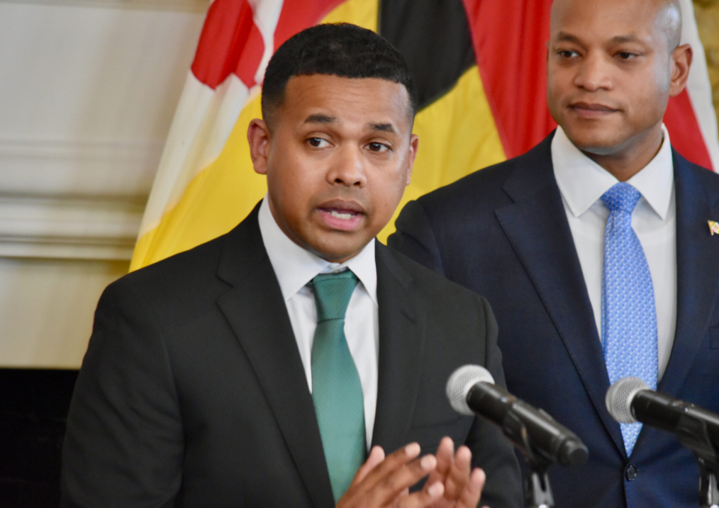 The image features two men standing at a podium. In the background of the image is a beige wall with crown molding and a standing Maryland State flag. The man at the center of the image is Paul Monteiro Jr. He is a brown-skinned man with closely cropped black hair. He is wearing a dark colored suit jacket, a white collared shirt, and a green tie. He is gesturing with his hands and talking into two microphones placed in front of him. Behind Paul Montiero Jr. is another man. This man is a brown-skinned man who is bald. He is wearing a dark blue suit, a white collared shirt, and a light blue patterned tie. On the left lapel of his suit is a flag pin. He is staring at Paul Montiero. 