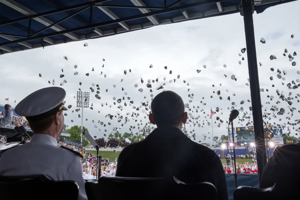 A man with a light skin tone in a white hat and suit sits besides President Obama in an stadium during the day. They watch white hats fly in the air 