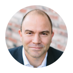 Ben Rhodes has a closed-lip smile. He has a light skin tone and thin brown hair. He is wearing a blue suit and white collared shirt.