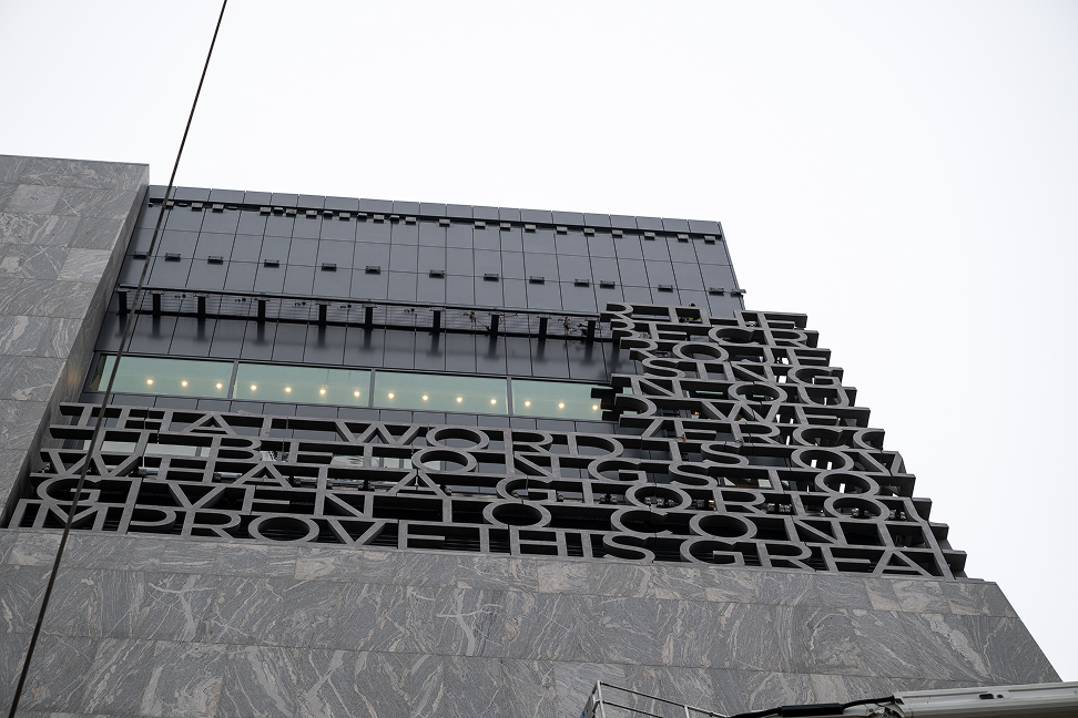 An upward shot of the installation of the “You Are America” letters on the side of the Obama Presidential Center Museum Building. It is gray outside. 