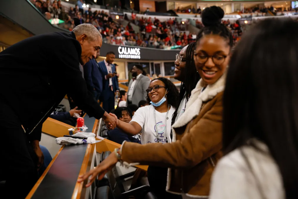 President Obama shakes hands with a young girl in a white tshirt and glasses in a packed basketball arena. Other young girls surround him smiling and talking to one another.
