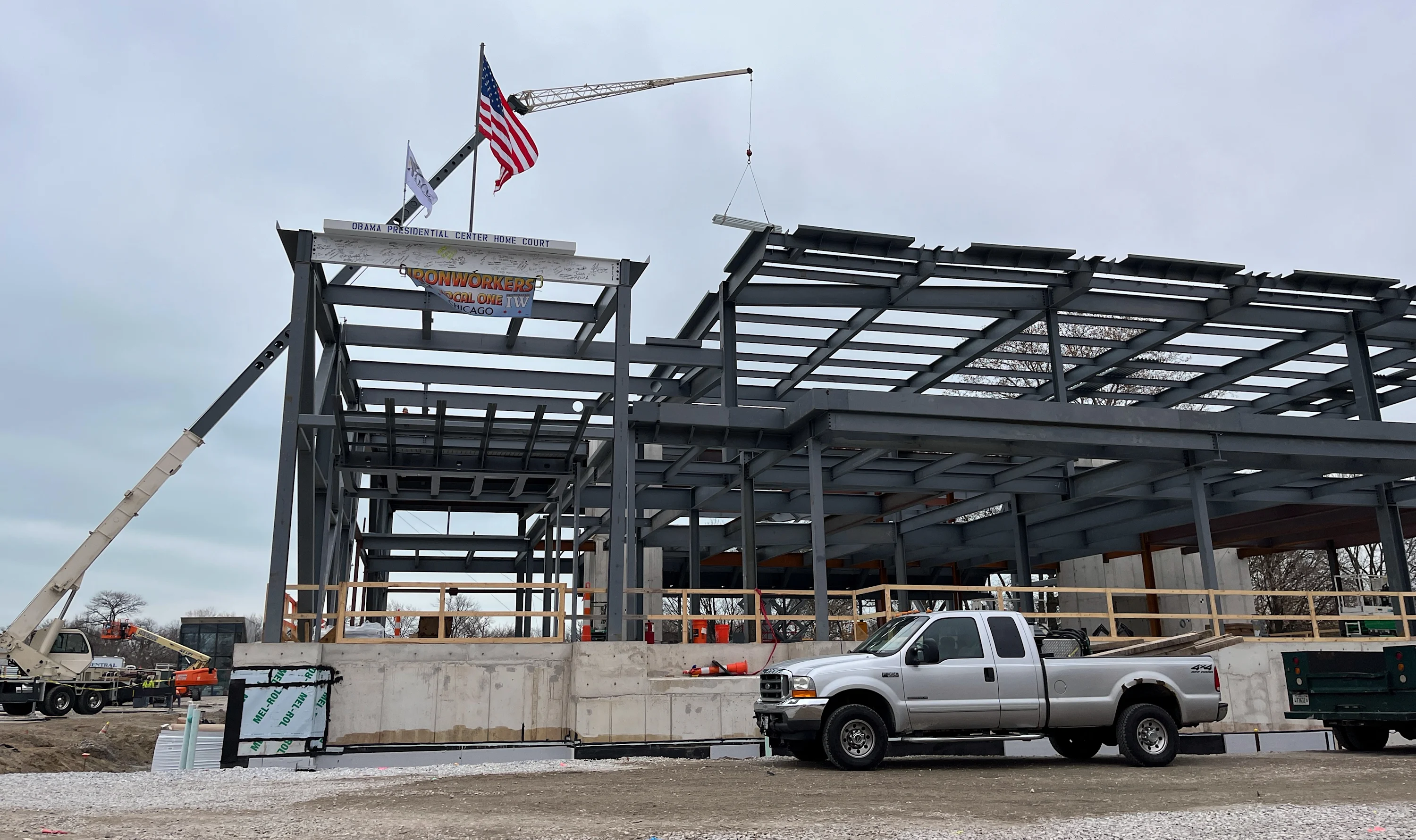  A silver pickup truck is parked in front of steel beams forming the shape of a large building. At the top is an American flag blowing lightly in the wind. A cloudy sky  and a large crane can be seen behind the building.