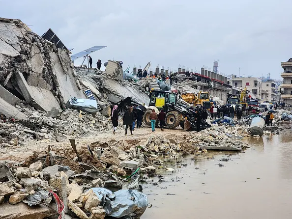 A landscape scene shows severe damage, with concrete road and bridge structures in massive heaps above a narrow pathway cleared in the rubble. A group of people of varying skin tones are seen at a distance wearing headscarves and assorted clothing near a small bulldozer. A body of brown water is shown on the right with rubble and debris along the shore.