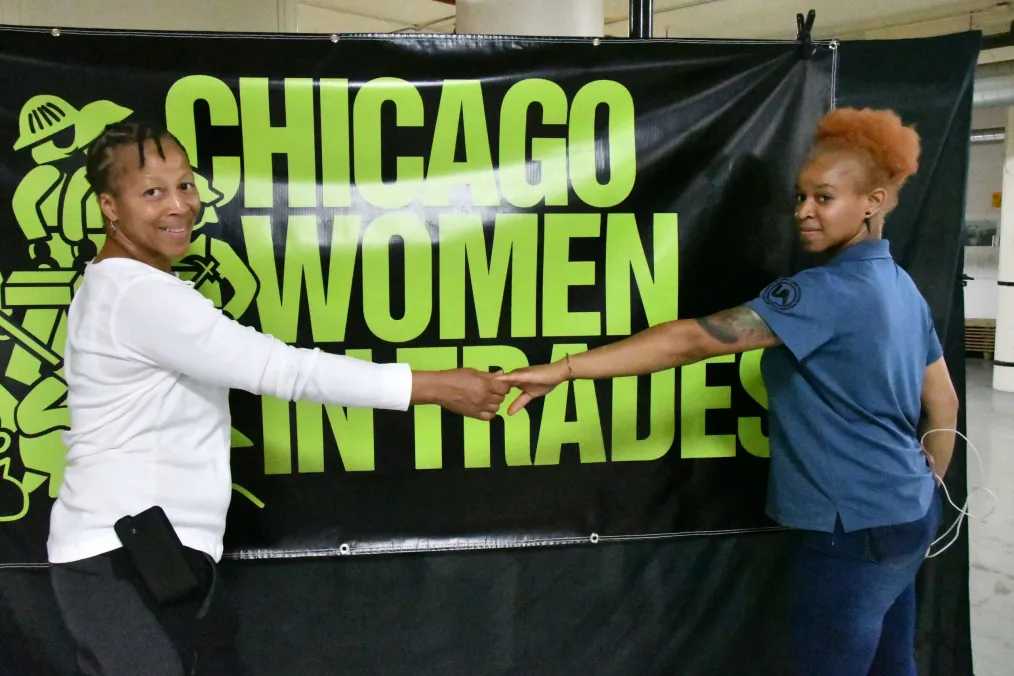 Zahrah Hill and her mom, Renee, hold hands and smile in front of a sign that reads "Chicago Women in Trades."