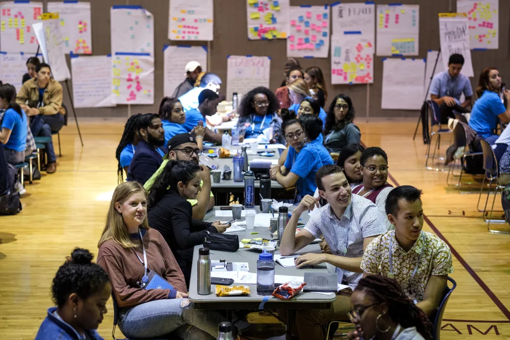 A group of individuals with a variety of skin tones sit at long gray tables with a brown wall with large white notepad paper and highlighted texts and sticky notes on it blurred in the background.